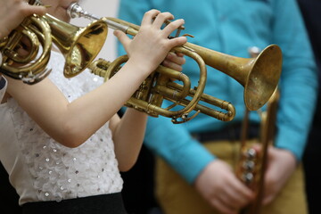Obraz premium An elegant child girl in a white sequined blouse plays a trumpet holding an old shabby musical instrument and pressing buttons with her fingers against the blurred background of an adult musician