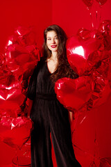 Valentine's Day. Beauty girl in black with red heart-shaped balloons having fun, on a red background