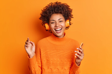 People entertainment and hobby concept. Cheerful young African American woman with curly hair holds modern smartphone listens music via stereo headphones poses against vivid orange background