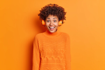 Fototapeta na wymiar Happy reaction concept. Overjoyed curly haired African American woman wears stereo headphones enjoys listening music dressed in casual jumper isolated over orange background. What wonderful sound