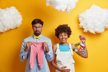 Diverse couple expect baby pose with toys and clothes for child going to become parents. Happy...