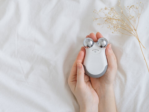 Microcurrent concept. Face massager with micro currents for home use and skincare on white bed. Female hands holds device for anti-aging and face lifting. Top view or flat lay, copy space for text.