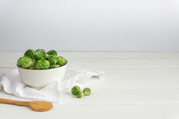 Frozen Brussels sprouts in a white bowl with a wooden spoon on white table, copy space