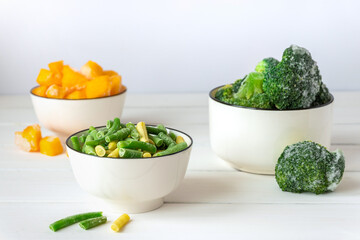 Frozen vegetables assortment. Frozen broccoli, French beans and pumpkin in white bowls on white table