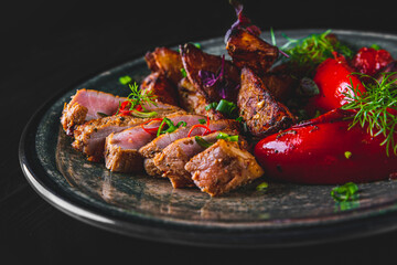 fried meat with potatoes, pepper, tomatoes, herbs and spices in plate on black wooden table background