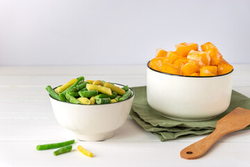 Frozen vegetables assortment. Frozen French beans and pieces of pumpkin in white bowls on white table