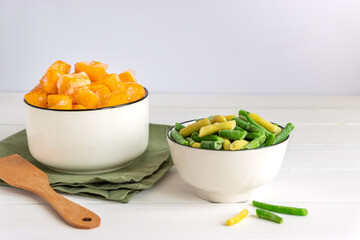 Frozen vegetables assortment. Frozen French beans and pieces of pumpkin in white bowls on white...