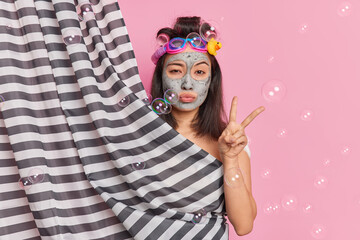 Serious brunette Asian woman makes peace gesture hides naked body behind shower curtain poses in douche applies clay mask for skin refreshment wears hair curlers prepares for special occasion