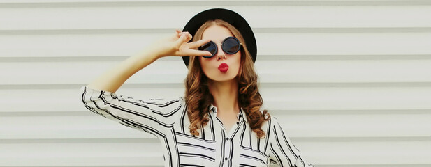 Portrait of stylish young woman blowing her red lips sending air kiss wearing a black round hat, shirt on a white background