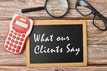 Top view of a calculator, spectacles and magnifying glass with written WHAT OUR CLIENTS SAY on wooden background. Selective focus.