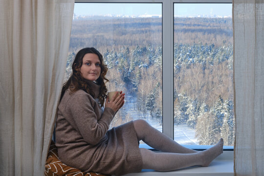 An adult woman sits at the window with a winter forest behind glass.