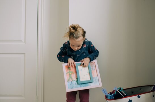 blonde girl holding onto paintings in frames in her bedroom smiling