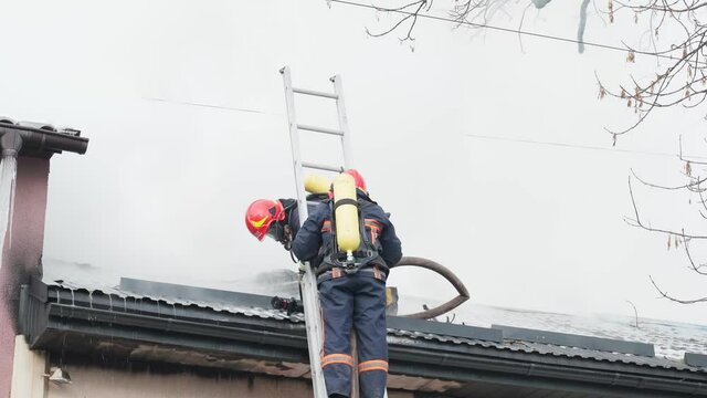Two firemen extinguish fire with hose on roof.