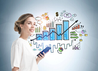 Attractive smiling businesswoman in formal wear holding a phone and standing near light blue wall with creative colorful bar chart sketch symbolizing business development process.