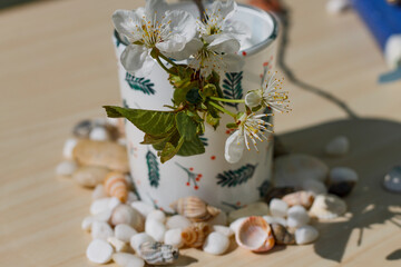 White candle with patterns on a beige table with sea stones, shells and a flowering cherry branch on a sunny day side view