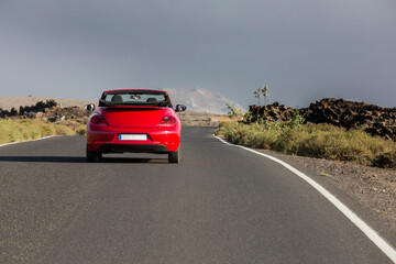 Red cabrio car driving empty paved road in desert volcanic landscape