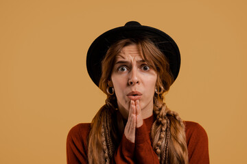 Photo of cute female farmer looks nervous and prays for something. White woman wears denim overall and hat isolated brown color background