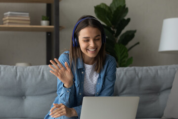 Smiling teenage female pupil sit on sofa in headphones wave hand to laptop screen greet teacher tutor at lesson consultation. Happy young woman say hi hello to friend speaking from home by video call © fizkes