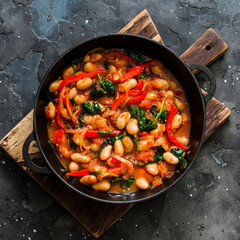 Vegetarian greek style tomato sauce, spinach, paprika, beans stew in a cast iron pan on a rustic board on a dark background, top view
