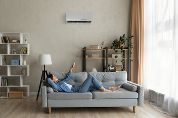 Glad millennial female buyer of air conditioner relax on sofa hold controller breath cool fresh air despite of hot summer day outside. Happy young lady regulate climate at home using modern ac device - obrazy, fototapety, plakaty