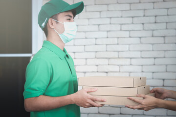 Young Asian delivery man in green T-shirt uniform and cap wearing face mask to prevent covid19 infection, holding pizza boxes and giving the boxes to customer hands, social distancing and new normal s