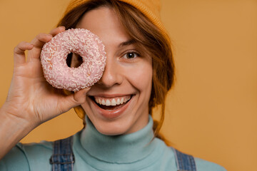 Close up photo of cute female farmer covers her eye by donut and smile. Wears denim overalls and hat, isolated brown color background