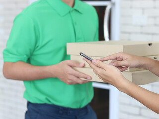 Delivery man in green T-shirt uniform and cap holding pizza boxes and giving to customer hands who pay with mobile phone, social distancing and new normal 