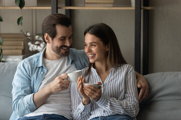 Happy married couple relax on cozy sofa at home cuddle discuss good news drink aromatic tea strong...