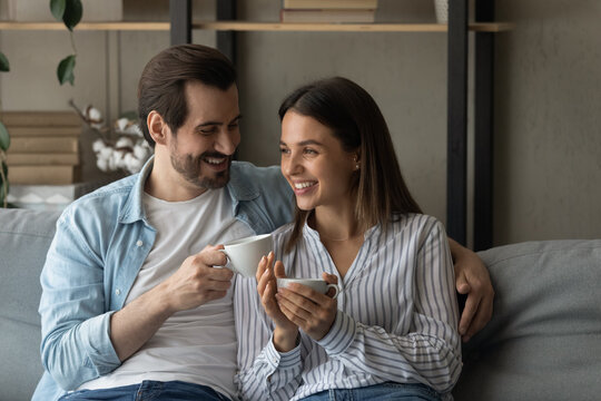 Happy married couple relax on cozy sofa at home cuddle discuss good news drink aromatic tea strong coffee. Smiling young man woman in love meet morning together sit on couch hold cups with hot drinks