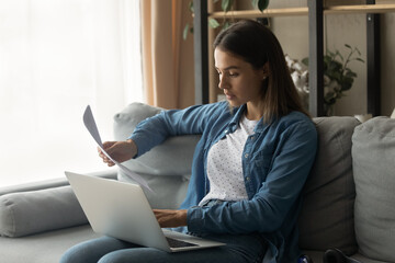 Serious millennial woman sit on couch hold laptop work with electronic document and paper hardcopy of loan insurance contract. Focused young female read official letter check actual information online