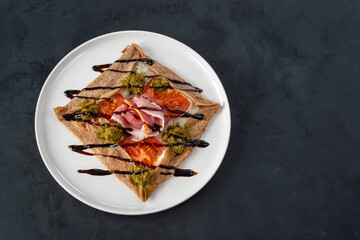 Galette bretonne with emmental cheese, homemade pesto, tomatoes and bacon on a black background.