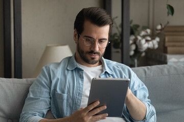 Serious millennial male in glasses sit on couch focus attention on digital pad screen read electronic book online. Concentrated guy student using tablet pc for distant learning watch education video