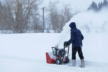 Heavy snowfall. A man using snowblower to clear driveway near residential house. Russia.