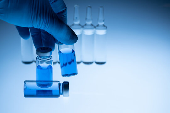 Hand Of Doctor With Medicine, Closeup. Vaccine Ampoules Are On The Table. The Background Is Illuminated From Below With Blue Light. Coronavirus And Influenza Vaccination Concept