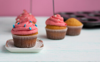 the muffin is on a saucer, decorated with cream and decor. Blurred cupcakes and kitchen utensils are in the background. The pink cream is applied in a spiral. Beautiful sweet dessert. 