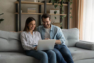 Happy husband wife newlyweds rest on sofa hold laptop choose furniture for new home buy consumer...