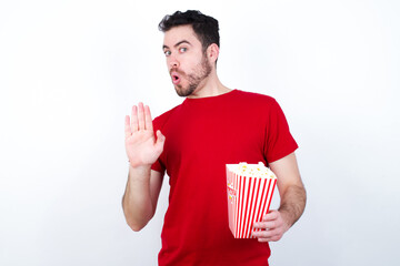 Young handsome man in red T-shirt against white background eating popcorn Moving away hands palms...