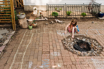 Little girl in arm cast draws on patio in backyard with sidewalk chalk