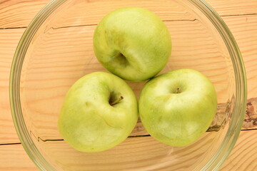 Three ripe green apples in a glass dish on a wooden table, close-up, top view.