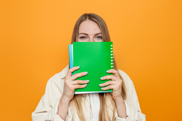 Close up portrait of a young student girl in in a beige jacket covers her face with a green exercise book isolated over orange background in studio