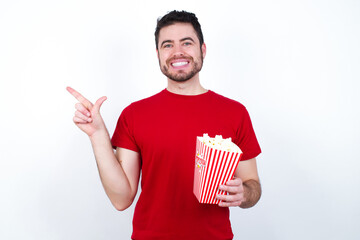 Young handsome man in red T-shirt against white background eating popcorn pointing up with fingers number eight in Chinese sign language BÄ.