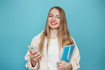 Blonde student girl holding mobile phone and notepad looking at the camera and smiling isolated on blue studio background. Online education