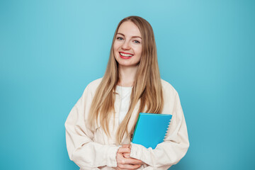 Blonde student girl holding a book, notebook smiling, looking at the camera isolated on blue background in studio