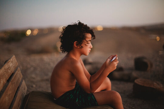 A young boy playing with his smartphone sitting on a bench in desert