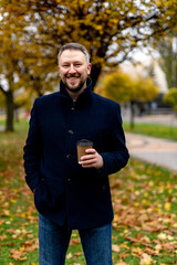 Obraz premium Smiling bearded male in formal clothes smiles to camera on autumn park background. Man with hot drinks in hand. Closeup.