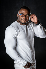 Cute pumped up black fashion model in total white outfit poses to the camera on dark studio background. Male in glasses. Closeup.