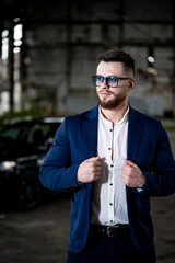 Fashion trendy man in blue suit poses on damaged warehouse interior. Male wears white shirt and modern glasses. Blurred background with black car.