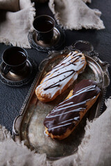 Chocolate and vanilla eclairs on an antique tray. Two cups of coffee in vintage dishes. Dark...