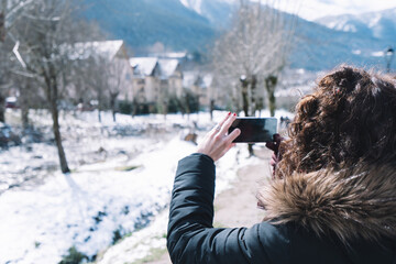 Beautiful curly brunette woman taking selfie portrait in the Pyrenees in winter, Spain. Mountains covered by snow, sunny day. girl video chatting on mobile phone sharing with friends family social