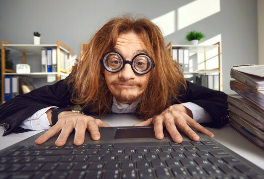 Funny nerd in round thick lens glasses sitting at desk and using laptop. Crazy looking office worker or computer geek typing on keyboard, searching for information on the Internet or doing accounts